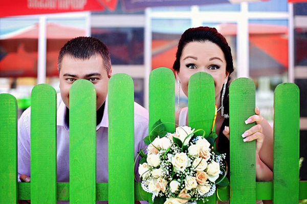 Bride and groom peeking over a bright green picket fence