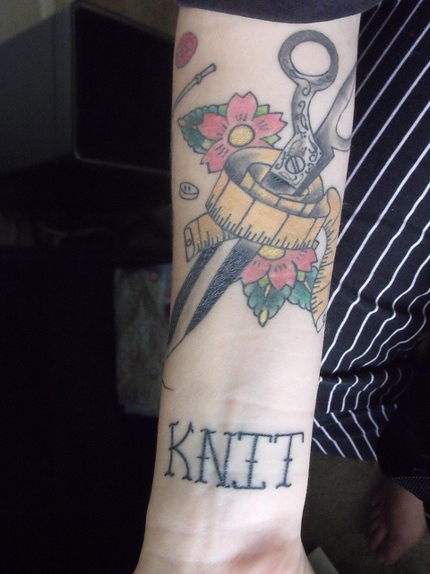 Forearm tattoo of ornate shears, measuring tape, flowers, and the word KNIT