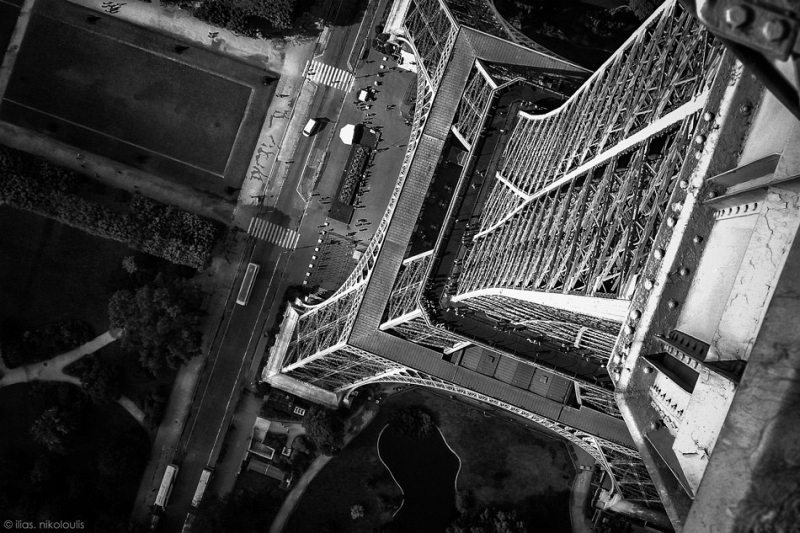 Black-and-white downward view from the Eiffel Tower ironwork to the park below