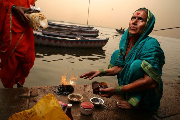 Woman in teal sari warming hands over a fire by boats