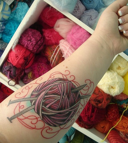 Forearm tattoo of a variegated yarn ball with knitting needles above bins of yarn